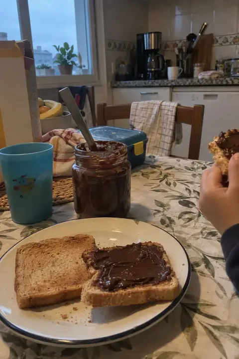 Tarro abierto de crema casera de cacao y avellanas con un cuchillo y tostadas en la mesa del desayuno